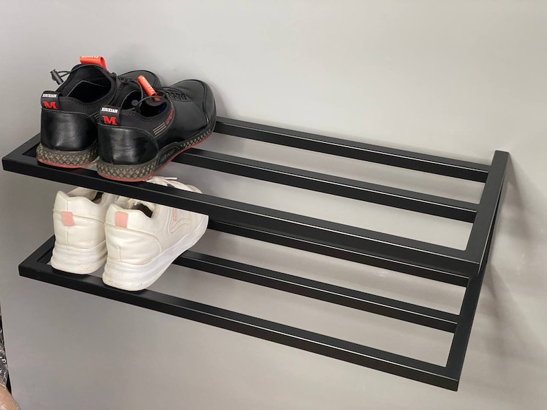 Może przedstawiać: Ścienny stojak na buty z czarnego metalu z dwoma poziomami. Na g&oacute;rnym poziomie znajduje się para czarnych sneakers&oacute;w z czerwonymi akcentami, a na dolnym para białych sneakers&oacute;w z jasnor&oacute;żowymi zapiętkami, o konstrukcji z poziomych listew.