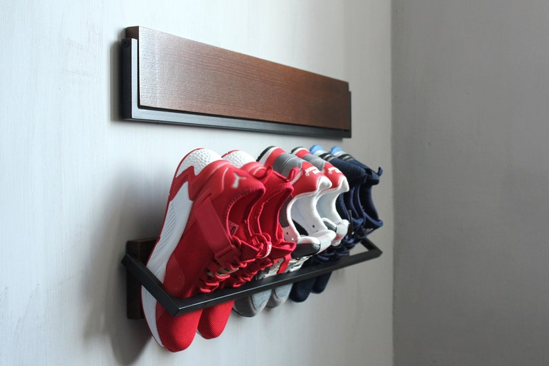 May include: A black metal shoe rack with six pairs of shoes, including red, white, and blue sneakers, mounted on a white wall with a wooden shelf above.