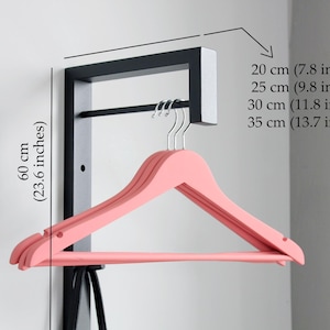 May include: A wall-mounted clothes rack with a black frame and a horizontal bar. It features a row of pink wooden hangers. The rack's dimensions are indicated: 60 cm (23.6 inches) high and widths of 20 cm (7.8 inches) to 35 cm (13.7 inches).