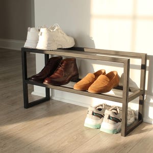 May include: A two-tier rectangular black metal shoe rack featuring a minimalist open frame design. The top shelf holds a pair of white trainers and a pair of brown leather boots. The bottom shelf holds a pair of tan loafers and white trainers.