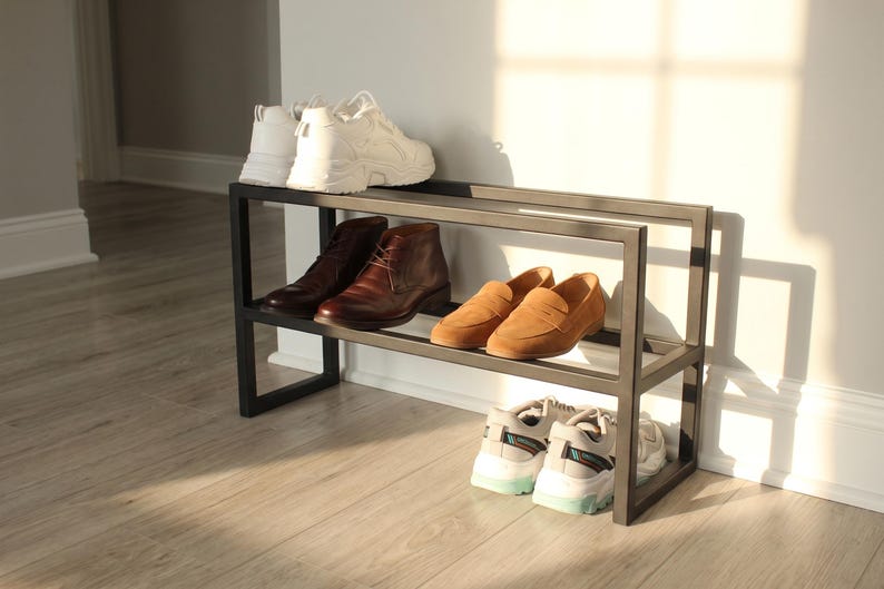 May include: A two-tier rectangular black metal shoe rack featuring a minimalist open frame design. The top shelf holds a pair of white trainers and a pair of brown leather boots. The bottom shelf holds a pair of tan loafers and white trainers.