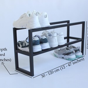 Op de afbeelding: Een zwart metalen schoenenrek met twee niveaus en witte en grijze sneakers op de planken. De tekst geeft een hoogte van 34 cm, een plankdiepte van 20 cm en een verstelbare breedte van 30 tot 120 cm aan voor dit opbergmeubel.