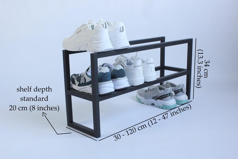 Op de afbeelding: Een zwart metalen schoenenrek met twee niveaus en witte en grijze sneakers op de planken. De tekst geeft een hoogte van 34 cm, een plankdiepte van 20 cm en een verstelbare breedte van 30 tot 120 cm aan voor dit opbergmeubel.
