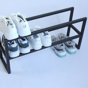 Op de afbeelding: Een zwart metalen schoenenrek met twee horizontale niveaus. Het bovenste niveau bevat twee witte sneakers, terwijl het onderste niveau twee witte sneakers met donkere accenten en twee grijze sneakers met lichtblauwe en zwarte zoolaccenten bevat.