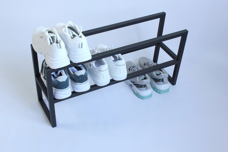 Op de afbeelding: Een zwart metalen schoenenrek met twee horizontale niveaus. Het bovenste niveau bevat twee witte sneakers, terwijl het onderste niveau twee witte sneakers met donkere accenten en twee grijze sneakers met lichtblauwe en zwarte zoolaccenten bevat.