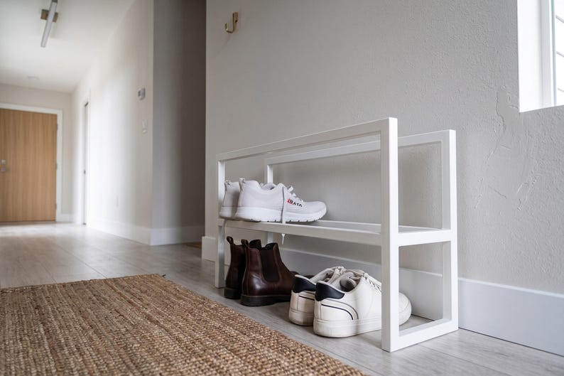 Op de afbeelding: Een wit metalen schoenenrek met twee niveaus en een rechthoekig frame. Op de bovenste plank staat een paar witte sportschoenen met rode accenten. Op de onderste plank staat een paar bruine leren enkellaarsjes en een paar witte sneakers.
