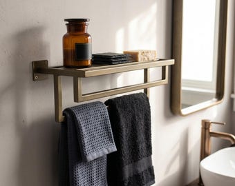 May include: A wall-mounted brass bathroom shelf with a rectangular frame and a lower bar for hanging towels. The shelf holds an amber glass jar, a stack of dark square coasters, and a bar of soap. Two textured towels, one grey and one black, hang below.