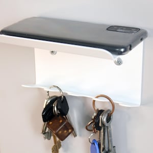 May include: White wall-mounted key holder with a shelf on top for a phone. The key holder has two hooks for keys and a black phone is on the shelf.