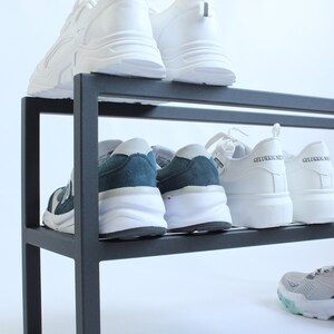 May include: A black metal shoe rack with two tiers holding several pairs of trainers. The top shelf features white trainers, while the middle shelf displays dark blue and white trainers alongside white trainers with the text GELUKKIG NIE.