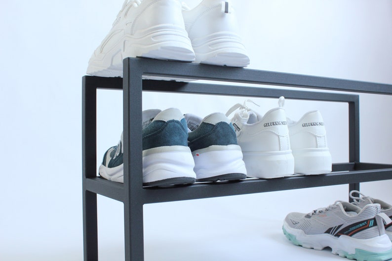 May include: A black metal shoe rack with two tiers holding several pairs of trainers. The top shelf features white trainers, while the middle shelf displays dark blue and white trainers alongside white trainers with the text GELUKKIG NIE.