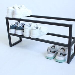 Op de afbeelding: Een zwart metalen schoenenrek met twee horizontale niveaus. Het bovenste niveau bevat een paar witte sneakers. Het middelste niveau bevat een paar donkergroene en witte sneakers en een paar witte sneakers. Onderaan staan grijze sneakers.