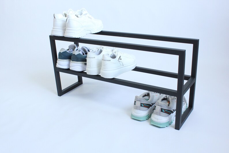 Op de afbeelding: Een zwart metalen schoenenrek met twee horizontale niveaus. Het bovenste niveau bevat een paar witte sneakers. Het middelste niveau bevat een paar donkergroene en witte sneakers en een paar witte sneakers. Onderaan staan grijze sneakers.