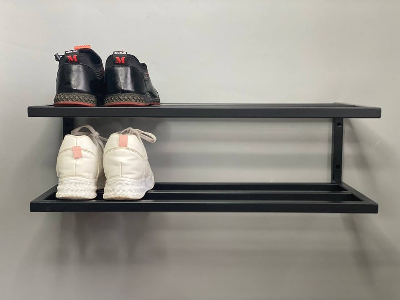 Może przedstawiać: Dwupoziomowy, ścienny stojak na buty wykonany z czarnego metalu. Na g&oacute;rnej p&oacute;łce znajduje się para czarnych sneakers&oacute;w z czerwonymi akcentami i literą M na pięcie. Na dolnej p&oacute;łce znajduje się para białych sneakers&oacute;w z jasnor&oacute;żowymi zapiętkami.