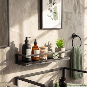 May include: A black metal wall-mounted shelf holding a matte black soap dispenser, an amber glass spray bottle, a clear jar with cotton swabs, a rolled green towel, and a small potted succulent plant against a textured gray wall.