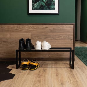 May include: A black metal shoe rack with a rectangular frame holding a pair of black high-top trainers and a pair of white low-top trainers on the top shelf, with a pair of yellow and black sandals placed on the floor beneath the rack.
