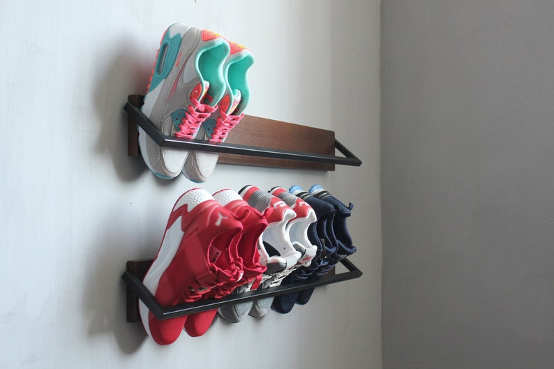 May include: A wall-mounted shoe rack with two shelves. The top shelf has two pairs of sneakers, one pair is white with pink and turquoise accents, the other pair is white with red and turquoise accents. The bottom shelf has six pairs of sneakers, two pairs are red, two pairs are white, and two pairs are navy blue.
