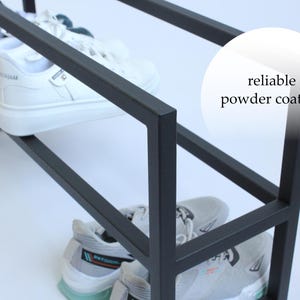 Op de afbeelding: Een zwart metalen schoenenrek met een rechthoekig frame en een matte afwerking. Het rek bevat verschillende paren sportschoenen op twee niveaus. Een cirkelvormige afbeelding aan de rechterkant bevat de tekst reliable powder coating.