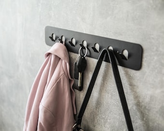 May include: A black metal wall-mounted hook rack with six hooks. A pink jacket, a black purse, and keys are hanging on the hooks.