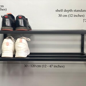 Może przedstawiać: Czarny, dwupoziomowy metalowy regał na buty montowany do ściany. Regał ma wysokość 20 cm, standardową głębokość 30 cm i szerokość od 30 do 120 cm. Para czarnych but&oacute;w sportowych znajduje się na g&oacute;rnej p&oacute;łce, a białe buty na dolnej p&oacute;łce.