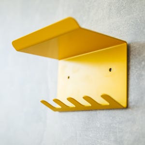 Könnte beinhalten: Ein leuchtend gelber Wandorganizer aus Metall mit einem Regal und vier gebogenen Haken. Er ist an einer grau strukturierten Wand befestigt. Das Design ist modern und minimalistisch.