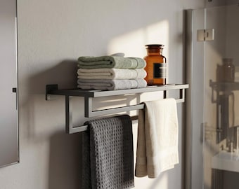 May include: A matte black metal wall-mounted bathroom towel rack featuring a top shelf holding a stack of three folded towels in sage green, cream, and grey, with a dark amber glass bottle and two additional towels hanging from the lower bar.