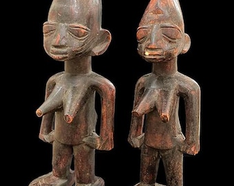 May include: Two antique wooden statues of human figures. The statues are carved from dark wood and have a weathered appearance. Each figure has a stylized face, arms at their sides, and large feet. The figures stand approximately 12 inches tall.
