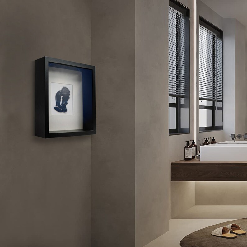 Pu&ograve; includere: Una cornice nera a scatola d'ombra con un passepartout bianco e un'immagine di silhouette blu all'interno. La cornice &egrave; appesa a una parete grigio chiaro.
