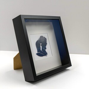 Pu&ograve; includere: Una scatola ombra incorniciata in nero con un passepartout bianco e una silhouette nera di due persone che si abbracciano. La scatola ombra ha uno sfondo blu.