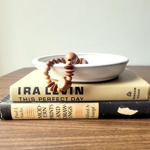 May include: A wooden bead necklace draped over a white ceramic bowl, resting on two vintage books. The books are titled "IRA LEVIN THIS PERFECT DAY" and "MODERN PRINTS AND DRAWINGS". The necklace features various shapes and shades of brown beads.