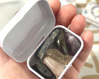 Puede incluir: Una pequeña lata de metal blanca con una tapa con bisagras, abierta para revelar una colección de piedras pulidas. Las piedras son de varios tonos de gris, marrón, negro y morado.