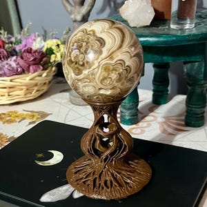 May include: A decorative sphere with swirling patterns in shades of brown and cream, displayed on a detailed brown stand. The stand has a root-like design. The sphere is on a black surface with a crescent moon and star design.
