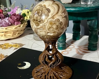 May include: A decorative sphere with swirling patterns in shades of brown and cream, displayed on a detailed brown stand. The stand has a root-like design. The sphere is on a black surface with a crescent moon and star design.