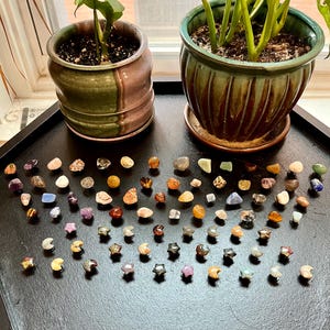 Pu&ograve; includere: Una collezione di piccole pietre e cristalli colorati, levigati, disposti a formare un motivo su una superficie nera. Le pietre sono di varie forme, tra cui stelle, lune e forme libere.