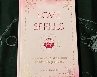 Puede incluir: Un libro rosa de tapa dura titulado "LOVE SPELLS" con detalles dorados. La portada presenta una ilustración de un frasco de poción con corazones y el texto "AN ENCHANTING SPELL BOOK OF POTIONS & RITUALS" de Minerva Radcliffe. El libro está sobre una tela verde oscuro.