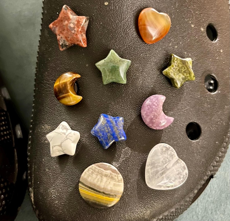 Pu&ograve; includere: Una collezione di 10 piccoli ciondoli in pietra preziosa lucida di varie forme e colori, tra cui stelle, cuori e lune crescenti. I ciondoli sono disposti su una superficie nera.