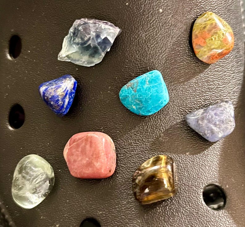 Pu&ograve; includere: Una collezione di otto piccole pietre levigate di vari colori, tra cui blu, verde, marrone, rosa e bianco. Le pietre sono disposte su una superficie marrone scuro.