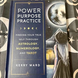 May include: A hardcover book titled "POWER PURPOSE PRACTICE" with gold lettering and a navy blue cover. The book's subtitle is "FINDING YOUR TRUE SELF THROUGH ASTROLOGY, NUMEROLOGY, AND TAROT" by Kerry Ward. A bookmark with a woman looking at a moon.