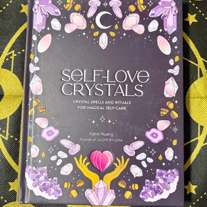 May include: A hardcover book titled "Self-Love Crystals" with a dark purple cover. The cover features crystal illustrations in shades of purple, pink, and white, along with a crescent moon and stars. The book's subtitle reads "Crystal Spells and Rituals for Magical Self-Care."