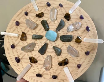 May include: A wooden, circular crystal grid with a flower of life design. The grid is adorned with various crystals, including clear quartz, amethyst, tiger's eye, and selenite wands. The crystals are arranged in a symmetrical pattern.