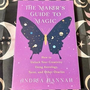 May include: A book titled "The Maker's Guide to Magic" with a purple cover. A butterfly design with celestial elements adorns the cover. The book's subtitle reads "How to Unlock Your Creativity Using Astrology, Tarot, and Other Oracles."