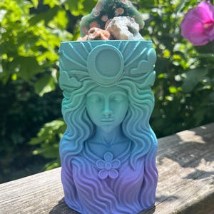 May include: A decorative, gradient-colored sculpture of a woman's face with long, flowing hair and a flower on the chest. The sculpture is in shades of blue and purple. The top of the sculpture holds a green and brown rock formation.