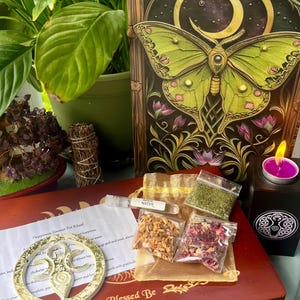 May include: A wooden box with a gold and white decorative circle, a small bag of dried herbs, a small bag of dried rose petals, a small bag of dried orange peel, a small vial of clear liquid labeled "Moon Water", and a small candle in a black holder. The box has the words "Blessed Be" printed on the front. The box is on a wooden surface.