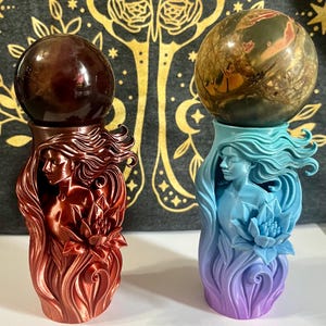 May include: Two decorative sculptures, each featuring a sphere atop a detailed base. One is a reddish-brown color, the other is blue and purple. The bases are sculpted with a woman's face and flowing hair, with a lotus flower.