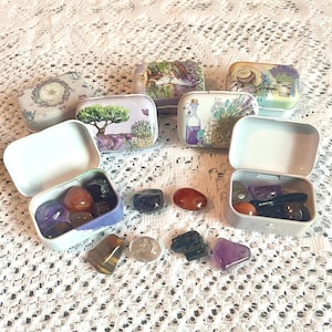 May include: A collection of small, decorative metal tins with various floral and landscape designs. Some tins are open, revealing colorful gemstones. The tins are arranged on a white, lace-like surface.