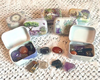 May include: A collection of small, decorative metal tins with various floral and landscape designs. Some tins are open, revealing colorful gemstones. The tins are arranged on a white, lace-like surface.