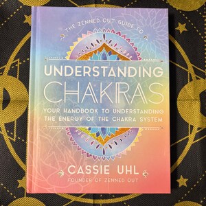 May include: A book titled "Understanding Chakras" with a colorful cover featuring a mandala design. The book's title is in large white letters, with the author's name, Cassie Uhl, and the phrase "Founder of Zenned Out" at the bottom.