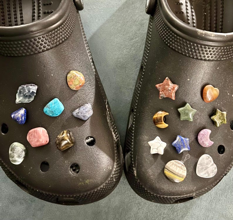 Pu&ograve; includere: Un paio di zoccoli neri con vari ciondoli di pietre preziose attaccati. I ciondoli includono stelle, lune, cuori e altre forme. I ciondoli sono di vari colori, tra cui rosso, blu, verde e marrone.