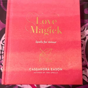 May include: A hardcover book titled "Love Magick" with gold lettering on a pink cover. The book includes the subtitle "Spells for Amour" and is authored by Cassandra Eason.
