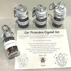 May include: Set of four small glass bottles with silver lids and keychains. Each bottle contains various crystals and has a label that reads "Car Protection". The bottles are arranged on a white card with the text "Car Protection Crystal Set" and a description of the crystals included in the set.