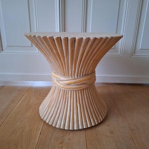 May include: A light beige, hourglass-shaped side table. The table is made of vertical, ridged wooden slats, with a woven band around the middle. The top is round and flat. The table is on a wooden floor.